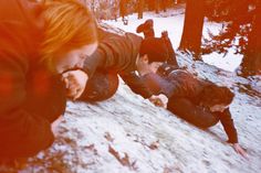 three people in the snow eating food and looking at something on the ground with trees behind them