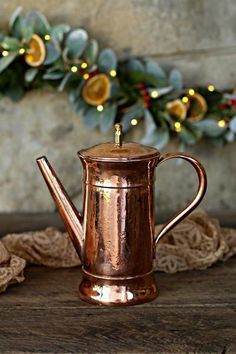 a copper coffee pot sitting on top of a wooden table next to a christmas wreath