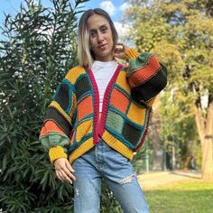 a woman wearing a multicolored knitted cardigan sweater and jeans standing in front of trees