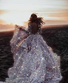 a woman in a long white dress standing on the beach at sunset with her back to the camera