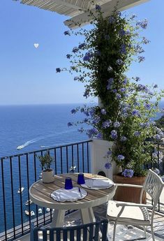 an outdoor table and chairs on a balcony overlooking the ocean with blue flowers growing over it