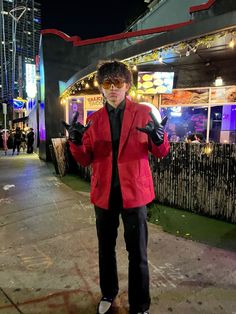 a man in a red jacket and black pants is standing on the sidewalk at night