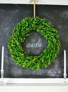 a wreath with the word noel written on it hanging from a chalkboard in front of candles