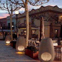 an outdoor dining area at dusk with tables and umbrellas in the foreground, lights on