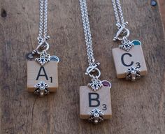 three necklaces with letters and charms hanging from them on a wooden surface, one has an initial charm