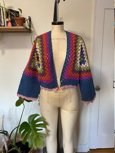 a mannequin wearing a multicolored crocheted sweater and shorts with a plant in the corner