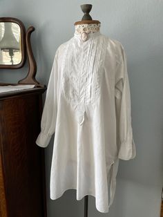 Beautiful, fine, cotton collarless French tunic from the early 1900s. Gorgeous front pleated detailnote the "M" monogram stitched on the front tab.   No buttons - made to wear with shirt studs and length longer at back with splits at the sides. Measures 16" from one top shoulder seam to the other with sleeve length 21".  Front length 31 " from top of collar with back length 37".  Would most likely fit woman's size medium to large. Tiny mends on the back and sleeve cuff placket but in excellent condition for at least 100 years old. A lovely creamy white, light summer shirt that could be worn as a tunic with leggings, nightshirt, or even a casual loose dress. Cameo brooch not included. Button-up Tunic For Daywear, Cotton Tunic Blouse With Buttons, Vintage Cotton Top With Stand Collar, Long Sleeve Tunic With Buttons For Daywear, Classic Cotton Blouse With Covered Buttons, Cotton Tops With Pintucks For Daywear, Button-up Daywear Blouse With Pintucks, Daywear Button-up Blouse With Pintucks, Button-up Blouse With Pintucks For Daywear