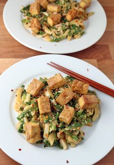 two white plates topped with tofu and vegetables next to chopsticks on a wooden table