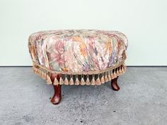 an upholstered foot stool with tassels on the legs and back rests against a wall