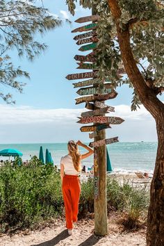 a woman standing next to a wooden pole with many signs on it near the beach