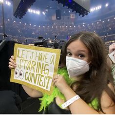 a woman wearing a face mask holds up a sign that reads let's have a staring contest