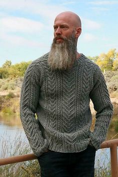 a man with a long beard wearing a sweater and jeans stands by the water in front of a fence