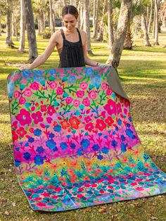 a woman standing next to a colorful blanket in the grass with trees behind her and holding it up