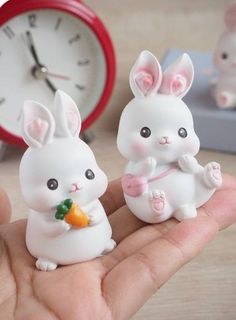 two small white rabbits holding carrots in front of a red and white alarm clock