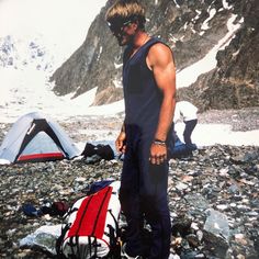 a man standing on top of a rocky beach next to a tent and snow covered mountains