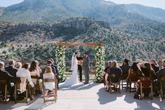 Captured atop a wooden platform in Mount Charleston, this wedding sets a breathtaking scene with the bride's timeless wedding dress cascading down steps under a rustic arbor of white blooms and green foliage. Guests, seated in rustic wooden chairs, watch as the couple shares their intimate moment against a backdrop of rugged mountains and clear blue skies. 🌼✨ #weddingideas #weddingdress #weddinghairstyles #mountcharlestonwedding #outdoorweddings Rustic Arbor, Rugged Mountains, Wooden Chairs