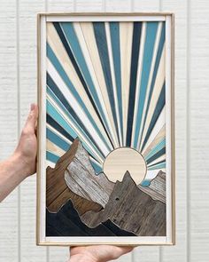 a person holding up a painting with mountains and sun in the background on a white brick wall