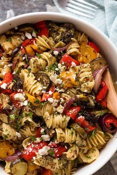 a white bowl filled with pasta and vegetables on top of a table next to a wooden spoon