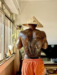 a tattooed man wearing an orange shorts and a straw hat stands in front of a window