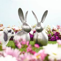 two metal rabbits sitting in the grass with flowers around them and one bunny is looking at it