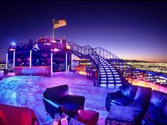 an outdoor lounge area with chairs and tables overlooking the city lights at night, lit up by colorful lighting