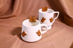 two white mugs with brown teddy bears on them sitting on a tan cloth covered surface