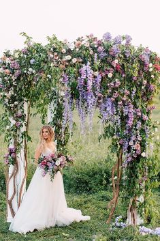 a woman in a wedding dress is standing under an arch with purple flowers on it