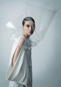 a woman wearing a white dress under a plastic covering her head and shoulders, standing in front of a gray background