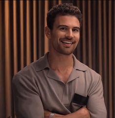 a smiling man with his arms crossed in front of a wooden wall and curtain behind him