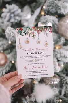 a person holding up a christmas party card in front of a tree with ornaments on it