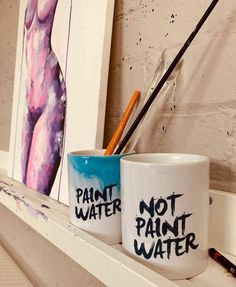 two coffee mugs with words painted on them sitting on a shelf in front of a painting