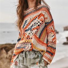 a woman standing on the beach wearing an orange and white crochet sweater with pockets