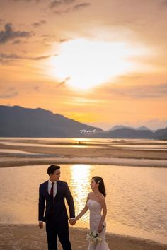 a bride and groom holding hands on the beach at sunset with mountains in the background