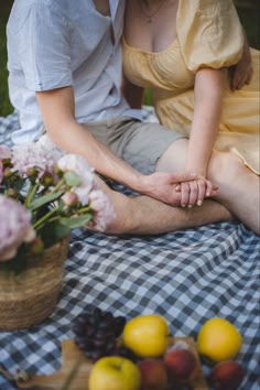 a man and woman sitting on a blanket with fruit in front of them, one holding the other's hand