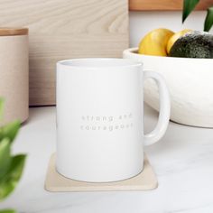 a white coffee mug sitting on top of a counter next to a bowl of fruit