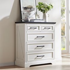 a white dresser with black top and drawers in front of a window next to a potted plant