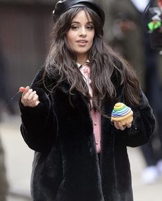 a woman in a black coat and hat is holding a cupcake while she walks down the street