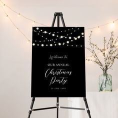 a black and gold christmas party welcome sign on an easel with lights in the background