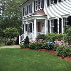 a white house with black shutters and flowers in the front yard on a sunny day