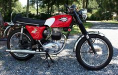 a red and white motorcycle parked next to a tree on gravel road with trees in the background