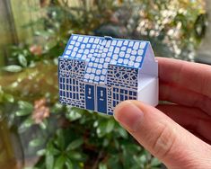 a hand holding up a small blue and white paper house in front of some plants