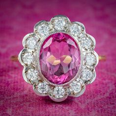 A spectacular Edwardian inspired daisy cluster ring bezel set with a vibrant oval cut pink topaz in the centre (approx. 5ct), haloed by twelve sparkling brilliant cut cubic zirconia petals. Pink topaz comes in shades of soft pink and fiery red and like a ruby is considered a stone of the heart. A symbol of femininity and romance, it's said to bring good fortune in love, providing happiness and vitality to any relationship. The ring is crafted in silver and the band is gilded in 18ct yellow gold. Classic Pink Cluster Ring With Halo Setting, Pink Round Cluster Ring With Halo Setting, Classic Pink Cluster Ring For Formal Occasions, Classic Pink Diamond Cluster Ring, Heirloom Pink Ring With Halo Setting, Pink Diamond Cluster Ring With Halo Setting, Classic Pink Halo Ring, Classic Pink Cluster Ring With Center Stone, Pink Cluster Ring With Gemstone