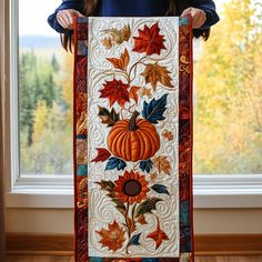 a woman holding up a quilt with autumn leaves and pumpkins on it in front of a window