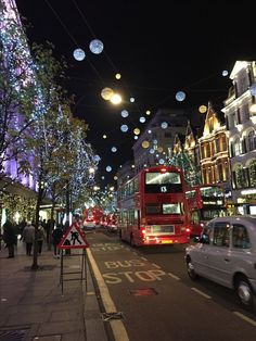 a red double decker bus driving down a street next to tall buildings and christmas lights