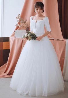 a woman standing in front of a pink curtain wearing a white dress and holding a bouquet