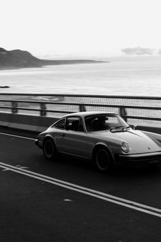 a black and white photo of a car driving on the road by the ocean with mountains in the background