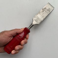 a hand holding a red handled knife on top of a white table next to a gray wall