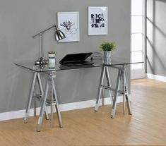 a glass desk with two metal legs and a laptop on top, in front of a gray wall