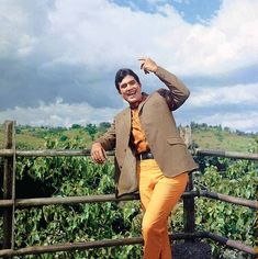 a man in yellow pants and a blazer leaning on a wooden fence with his hand up