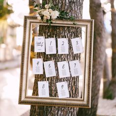 a tree with seating cards attached to it and flowers on the top, hanging from a gold frame